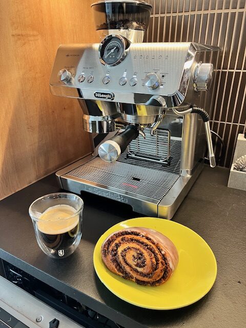 ekspres kolbowy De'Longhi La Specialista Opera na blacie kuchennym wraz ze szklanką zaparzonej kawy i drożdżówką na talerzyku